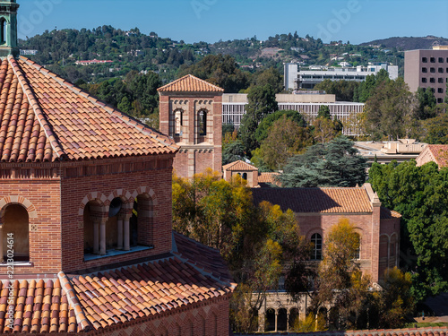 Romanesque revival brick architecture of UCLA campus shows tile roofs and bell towers framed by forested hills of Los Angeles under clear daytime skies.