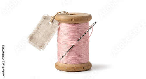 Vintage wooden spool with pink sewing thread, a sharp metal needle, and a small frayed beige linen tag attached by a string, isolated on a white background