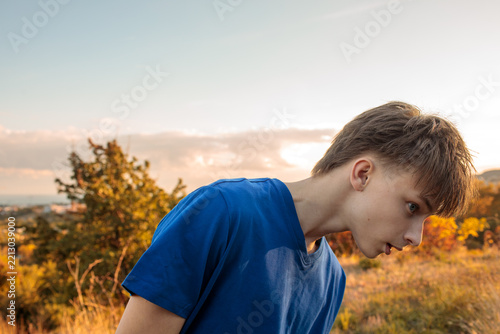 Teenager in a blue t-shirt and light jeans stands on a grassy hill at golden hour, dry autumn grass and orange bushes around, city and sea far behind, grainy film texture, high quality photo