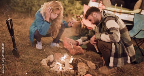 Couple cooks food by campfire in the woods while enjoying a night under the stars with friends around the car and tents in the background