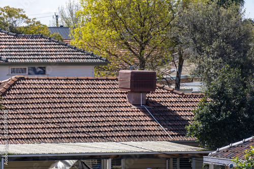 A terracotta tiled roof on a suburban Australian house, featuring an evaporative air cooling unit, framed by trees and neighbouring roofs. Home energy efficiency, residential climate control