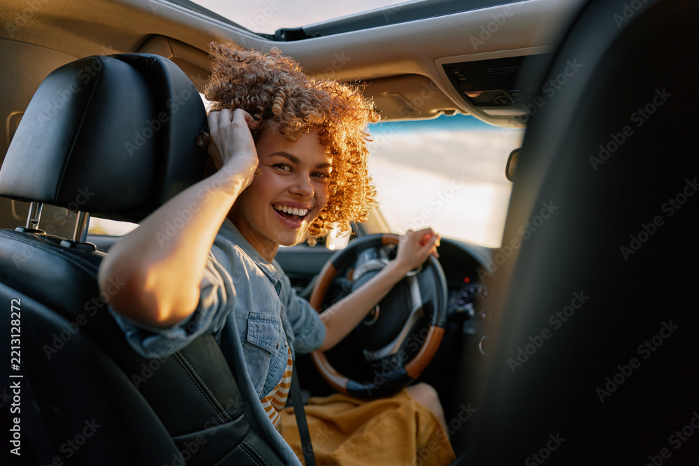 Obraz premium A young woman with curly hair smiles while driving a car in daylight, wearing a denim shirt and a seatbelt, capturing a bright casual moment on the road.