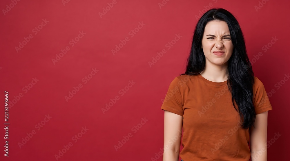 Fototapeta premium Portrait of a young woman with a disgusted facial expression wrinkling her nose against a red background with copy space