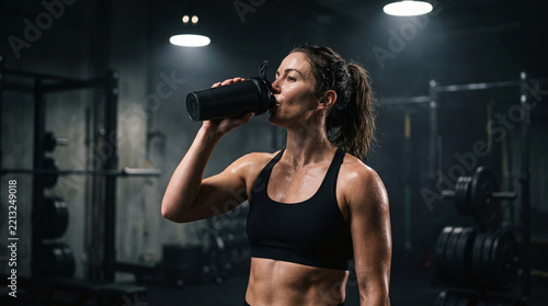 Sweaty female athlete drinking protein shake from shaker bottle in gym