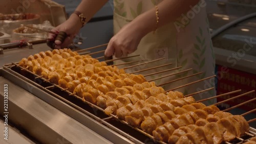 Spiced skewered gluten being grilled, with hands adjusting skewers on a commercial grill in a food stall.