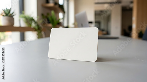 A blank white rectangular business card standing on a light grey office desk with blurred indoor background, featuring plants and workspace isolated on a transparent background