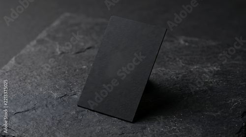 A single blank black business card standing upright on a rough dark grey stone surface with natural texture and soft lighting shadows isolated on a transparent background