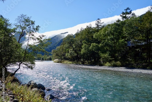 Scenic clear mountain river flowing through lush forest with mountain ridge engulfed in low clouds under blue sky in Kamikochi, Nagano, Japan