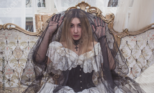 Mysterious gothic bride in a voluminous Victorian lace dress, black velvet corset and dramatic black veil at an ivy-covered old house. Dark romantic Halloween-inspired costume with antique atmosphere