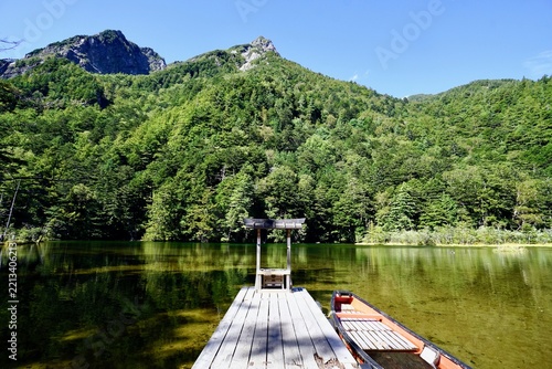 Serene alpine pond with wooden pier, small shrine torii gate, and traditional boat under towering mountains in Kamikochi, Nagano, Japan