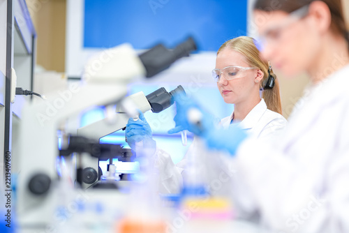 Blonde female scientist pipetting at a microscope while a brunette colleague works in the foreground during a microbial assay for an AGI-guided pathogen detection study