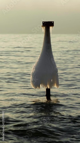 frozen metal pole in sea covered with ice structure vertical view, ice coated navigation post with icicles in ocean water, vertical view of frozen post with ice skirt on calm sea surface