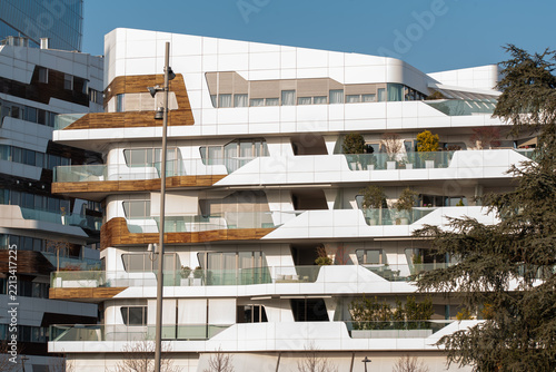 A contemporary residential building features sculptural white terraces, warm wood accents, and broad glass windows, creating a striking contrast against the clear blue sky and neighboring urban archit
