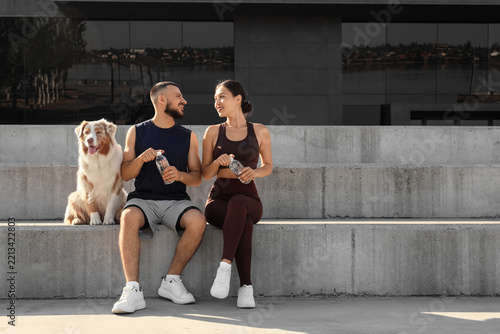 Healthy lifestyle and fitness, young couple with dog resting after workout and drinking water on concrete steps in urban environment