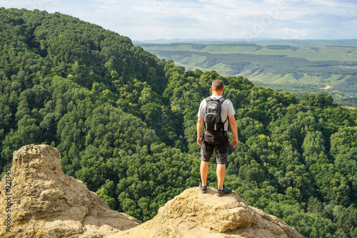Man tourist with a backpack standing on a rocky, protruding ledge against scenic green hills and wooded slopes