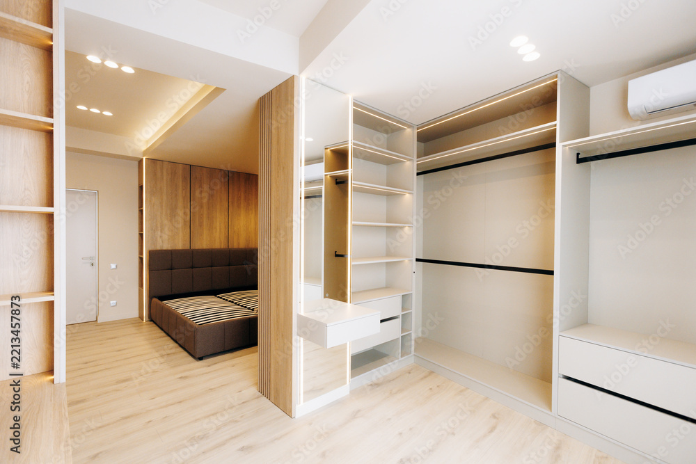 Fototapeta premium Bright modern walk-in closet interior with open shelving drawers and hanging rails, integrated vanity with mirror, connected bedroom view, neutral tones and recessed ceiling lights