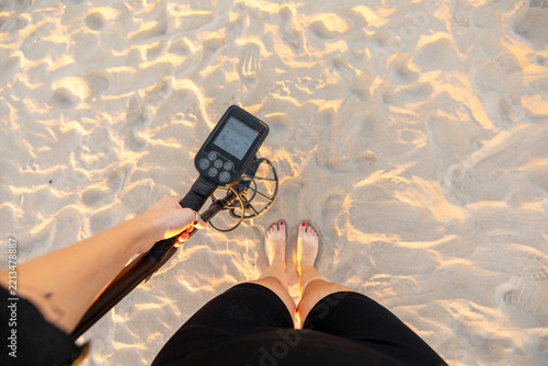 Top view of woman barefoot on sand holding metal detector at sunset