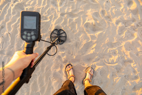 Man holding metal detector on sandy beach wearing sandals at sunset
