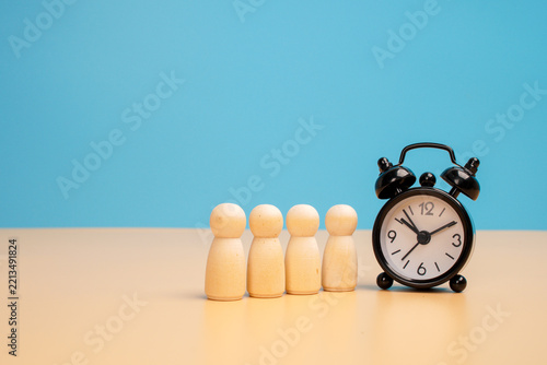 Fotografie Four wooden human figures standing in a row beside a black alarm clock on a beige surface against a blue background