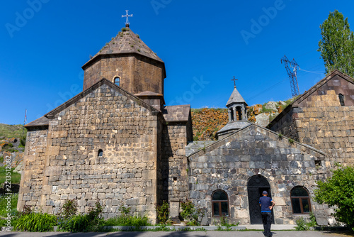Man wearing a cap walks along a stone pathway toward the arched entrance of Vanevan Monastery past ancient stone buildings and a carved khachkar, Artsvanist, Armenia