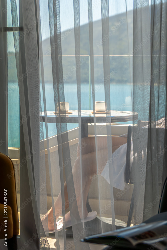 Naklejka premium Woman in a white bathrobe relaxing on a private hotel balcony overlooking the blue sea and mountains. View through sheer curtains creates a peaceful morning vacation and wellness atmosphere.