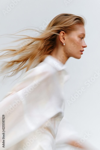 Elegant blonde woman in profile with windblown hair and flowing white clothing, captured with subtle motion blur on a minimalist background