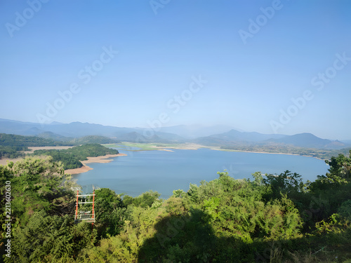 A tranquil reservoir stretches toward distant mountain ranges under a clear blue sky, framed by lush foreground greenery and a prominent electrical transmission tower overlooking the water.