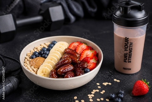 Healthy fitness oatmeal bowl with dates banana strawberry blueberry peanut butter and protein shaker on dark background