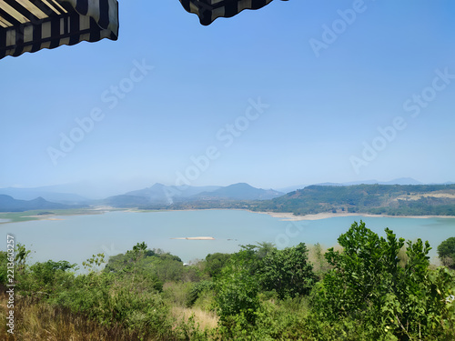 A tranquil vista overlooking a wide, calm reservoir surrounded by rolling hills and lush green vegetation, captured from a shaded vantage point on a bright, sunny day.