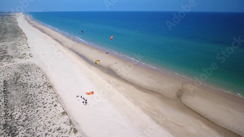 Incredible aerial view showing kitesurfers flying their colorful kites along a stunning sandy beach, with turquoise waves breaking on the shore during a beautiful sunny day