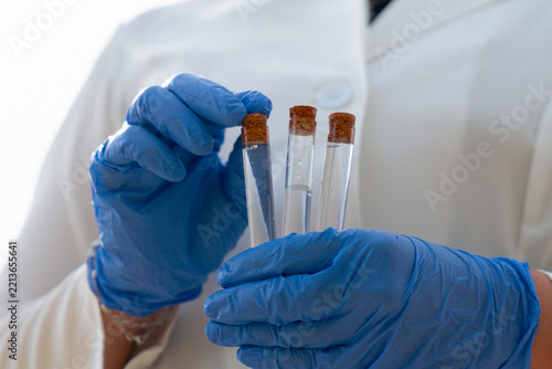 pneumonic plague outbreak scientist holding test tube sample, focused female epidemiologist in lab coat, face mask and blue gloves examining sample looking away from camera, forest and sky with copy s