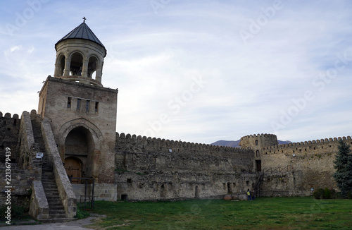 The stone belfry gatehouse and crenelated defensive walls enclose a grassy courtyard at the Svetitskhoveli Cathedral complex in Mtskheta, Georgia