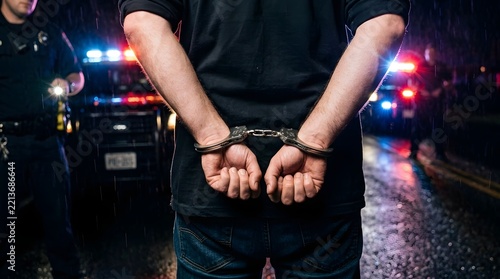 Close up of a man's hands in silver metal handcuffs being arrested by police officers at night on a wet street with flashing red and blue emergency vehicle lights in the background.