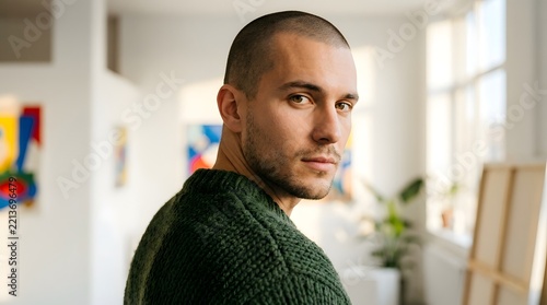 Portrait of a handsome young man with a short buzz cut and stubble wearing a textured green knitted sweater looking over his shoulder in a bright contemporary art gallery or studio setting.