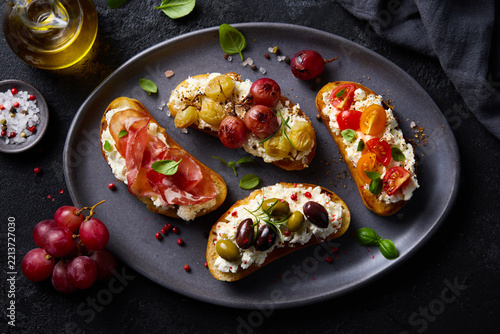 Italian crostini appetizers with prosciutto, roasted tomatoes, olives and ricotta. Dark grey background. Close up. Top view.