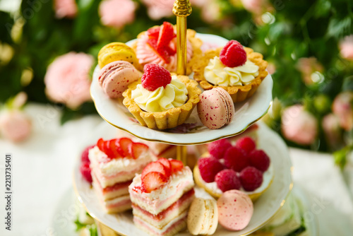 Afternoon tea sweet pastries, berry tartlets and macarons on a tiered stand. Garden rose background. Close up.