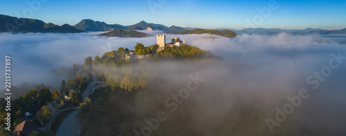 Fotografie Aerial drone view of the medieval Celje Castle rising above a sea of morning fog in Slovenia, with golden sunlight, forested hills and dramatic mountain scenery
