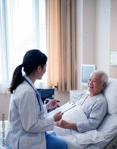 Caring Asian female physician listening to elderly male patient while taking medical notes in bright hospital ward