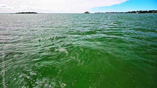 Point of view shot sailing across choppy green sea water with a distant island and coastline visible on the horizon under a cloudy sky