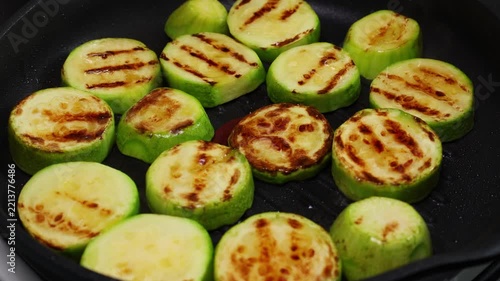 Round zucchini pieces grilled to perfection with visible char marks, cooking on a black grill pan. The zucchini slices present a mixture of lightly browned and green vibrant colors, showcasing a.