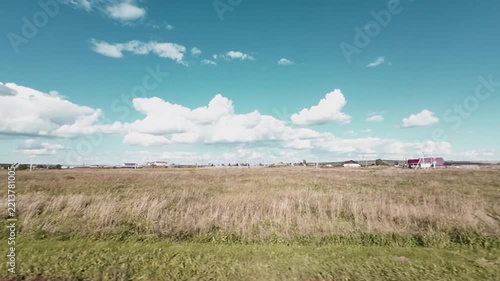 Vast open field covered with dry grass stretches toward the horizon, where small buildings and houses are visible in the distance, view from the car