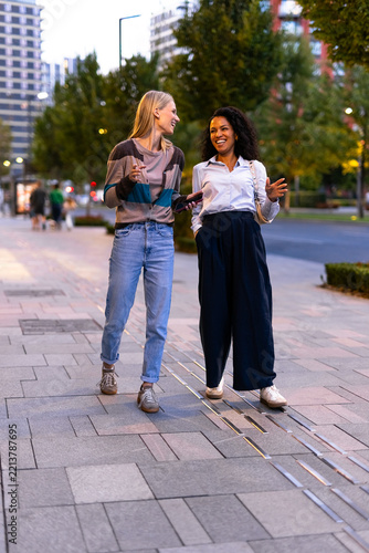 Two young women enjoying a conversation while walking urban streets, exploring commercial areas and interacting socially in the evening