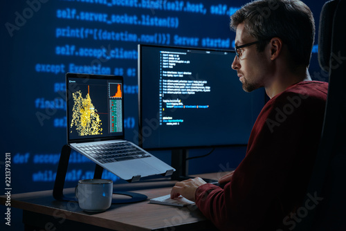 Developer sits in a dimly lit workspace, in front screens displaying code and data science data graph. Focused programmer working, deeply immersed in solving an artificial intelligence problem