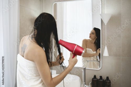 A woman stands in a bright, contemporary bathroom, holding a hair dryer and drying her wet hair. She is wrapped in a towel and gazes into the mirror, surrounded by minimalist decor