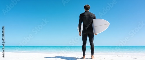 A lone surfer in a black wetsuit stands on a pristine white sand beach holding a surfboard while looking out at the turquoise ocean under a clear blue sky, ideal for travel and lifestyle advertising.