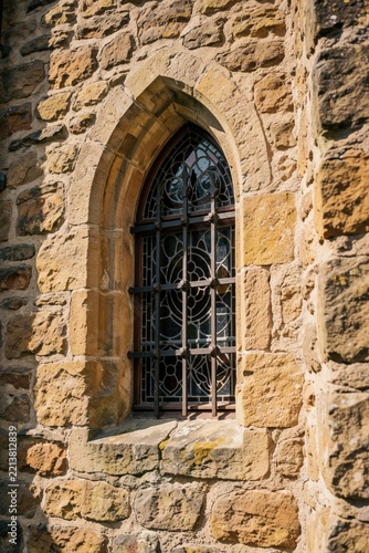 Close up of arched gothic window in old stone castle wall. Ancient medieval architecture features decorative iron grill, stained glass. Rustic masonry building exterior shows historic texture,