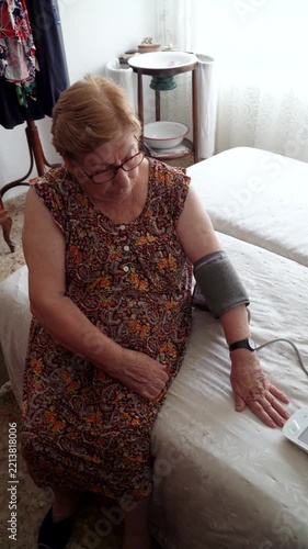 An elderly woman sitting on the bed, taking her blood pressure with an electronic blood pressure monitor