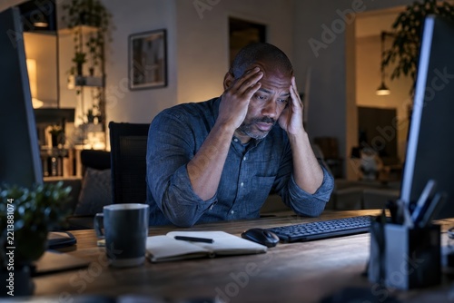 Exhausted man rubbing temples at desk facing computer screens