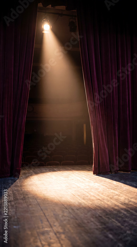 Empty theater stage framed by dark burgundy curtains with a warm spotlight shining across a wooden floor and rows of seats in the auditorium