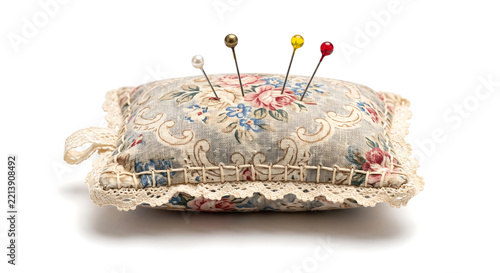 Rectangular hand-sewn pincushion made of vintage floral fabric with lace trim and four colorful sewing pins inserted on isolated on white background